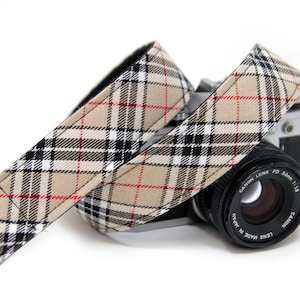 May include: A camera strap with a beige, black, and red plaid pattern. The strap is wrapped around a vintage Canon camera with a black lens. The strap's design is a classic plaid, adding a touch of style to the camera.