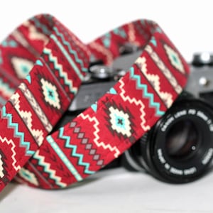 May include: A red, white, and blue patterned camera strap with geometric designs. The strap is wrapped around a black camera lens.