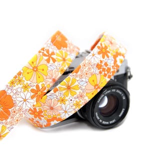 May include: A white camera strap with a floral pattern in shades of orange and yellow. The strap is wrapped around a black camera lens.
