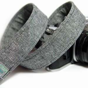 May include: A gray camera strap made of woven fabric with the text "PHAT STRAPS" and "HANDCRAFTED IN THE USA" printed on it. The strap is attached to a black camera with a silver lens.