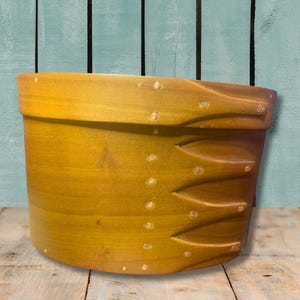 May include: A handcrafted, wooden, yellow-toned Shaker-style pantry box. The box features a rounded shape with a slightly angled rim and decorative dovetail joints. Small, white, evenly spaced pegs adorn the sides. The background is a weathered, blue-green wooden wall.