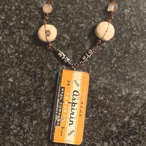 May include: A unique necklace featuring a vintage Aspirin box pendant. The necklace has a bronze-colored chain with cream-colored beads and two large, round, off-white beads. The pendant is rectangular, with the text "Aspirin" and other details.