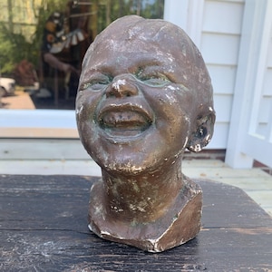 May include: A weathered bronze-toned sculpture of a laughing baby's head. The sculpture has visible texture and a distressed appearance, with white accents on the base. The head is positioned on a dark wooden surface.