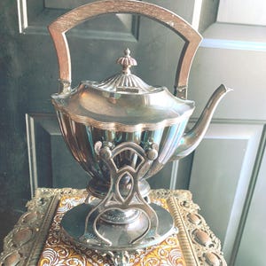 May include: A silver-colored tea kettle with a wooden handle and a decorative base. The kettle has a spout and a lid with a finial. It sits on a decorative stand with a patterned tile insert, showcasing an antique design.