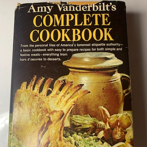 May include: A vintage cookbook titled "Amy Vanderbilt's Complete Cookbook" with a photo of a roasted leg of lamb on a bed of green beans in a copper pot. The book cover is black with gold lettering.