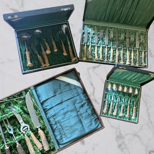 May include: Four antique cutlery sets in open black boxes with green velvet lining. The silverware features ornate handles with a light brown and cream color. The sets include knives, forks, spoons, and serving utensils.