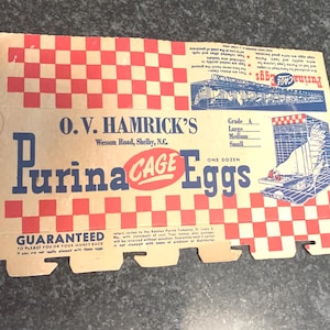 May include: Vintage Purina Cage Eggs carton with red and white checkered pattern. The carton features text including "O.V. Hamrick's" and "Purina Cage Eggs". A blue and white illustration of a chicken coop is also visible.