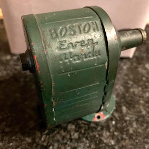 May include: Vintage green metal pencil sharpener with the text "BOSTON Ever-Hardy". The sharpener has a cylindrical body, a handle, and a base. The paint is chipped, revealing red primer.