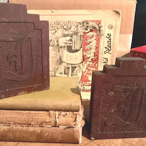May include: Two antique, dark brown bookends with a textured surface and a stylized letter "D" design. The bookends are displayed with a stack of old books and vintage ephemera, suggesting a theme of classic literature and vintage decor.