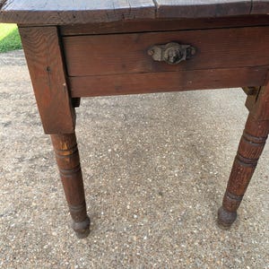 May include: A rustic, antique wooden table with a dark brown finish. The table features a single drawer with a decorative metal handle. The legs are turned and have a ball foot design. The tabletop is made of several wooden planks.