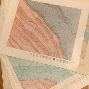 May include: A collection of vintage geological maps. The maps feature detailed illustrations with a color palette of pink, orange, blue, and gray. The maps are layered on top of each other, with the top map centered and the others partially visible.
