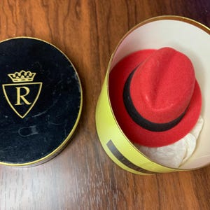 May include: A red fedora hat with a black band inside a yellow and white hat box. The box is open, revealing the hat and a white tissue paper. The box lid is black with a gold emblem.