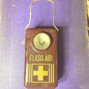 May include: A vintage brown metal flashlight with a handle. The flashlight has a round lens and the words "FLASH-AID" above a gold cross symbol. The flashlight is a rectangular shape.