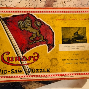 May include: Vintage Cunard Line jigsaw puzzle box. The box features a red flag with a gold lion emblem, the Cunard logo, and an image of the ship "Berengaria." The puzzle size is 16 x 12 inches and has about 150 pieces.