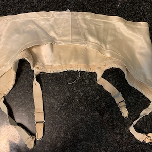 May include: A white satin garter belt with three elastic straps and metal clips. The garter belt has a ruffled edge and is in good condition.