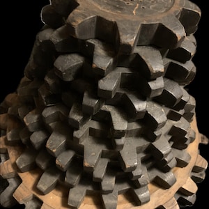 May include: A stacked wooden gear sculpture with a dark finish. The gears are arranged in a conical shape, with a central wooden dowel. The base gear is a lighter wood color. The sculpture is set against a black background.