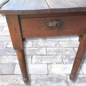 May include: A rustic, wooden table with a dark stained top and a single drawer. The drawer has a decorative metal handle. The table has four turned legs and shows signs of wear, giving it a vintage appearance.