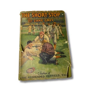 May include: A vintage hardcover book titled The Short-Stop by Zane Grey, featuring a color illustration of baseball players on a field. The cover shows a player being attended to on the grass, with text at the top and bottom edges.