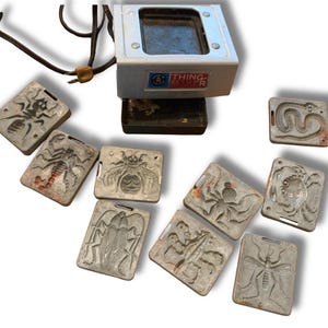 May include: A silver-colored Thingmaker heating unit with a black power cord and a collection of eight rectangular metal molds featuring embossed insect and arachnid designs including a scorpion, bee, spider, and beetle.