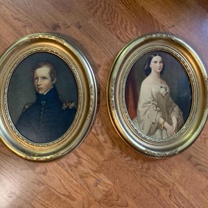 May include: Two oval portraits in ornate gold frames. One depicts a man in a dark blue coat, the other a woman in a light-colored dress. The portraits are set against a wooden background.
