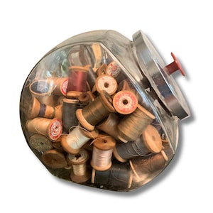 May include: A clear glass jar with a slanted opening and a silver lid featuring a red knob, filled with a collection of vintage wooden and plastic sewing thread spools in various colors including red, grey, brown, white, and tan.