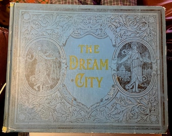 May include: A light blue hardcover book titled The Dream City with gold lettering. The cover features intricate embossed floral scrollwork and two circular illustrations of figures in classical robes, one standing by an easel and one by a building.
