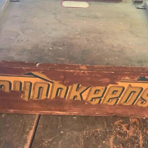 May include: A vintage wooden box with a hinged lid. The box is stained a dark brown with visible wear and tear. The word "Poughkeepsie" is painted in gold lettering on the side of the box, with some paint chipping.