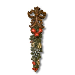 May include: A vertical wall hanging featuring a carved brown wooden bow at the top, cascading down into a cluster of painted fruit and foliage. The arrangement includes red apples, green leaves, striped melons, and blue grapes in a relief design.