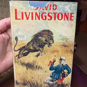 May include: Vintage book cover titled "David Livingstone" with a dramatic illustration of a lion attacking a man in a blue jacket. The cover is aged, with visible wear and tear, and the title is in large red letters.