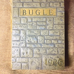 May include: A vintage yearbook titled "BUGLE" with a stone-like cover. The cover features a brick pattern, a gold-colored rectangular plaque with the title, and the year "1936" in gold lettering. The book is closed and resting on a textured surface.