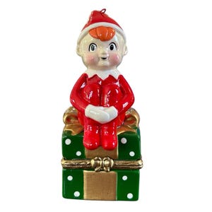 May include: A ceramic Christmas elf ornament. The elf is dressed in a red suit and hat with a white collar and cuffs. It sits atop a green gift box with white polka dots and gold accents. A festive holiday decoration.