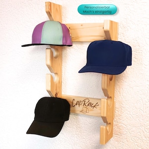 Könnte beinhalten: Ein Holz-Cap-Rack mit drei Baseballkappen: eine lila und grüne Kappe, eine marineblaue Kappe und eine schwarze Kappe. Auf dem Rack steht "Cap Rack" und eine Kappen-Grafik. Der Text "Personalisierbar Mach's einzigartig" ist ebenfalls sichtbar.