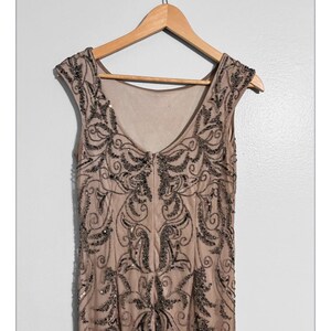 May include: A beige sleeveless dress with intricate black beadwork, hanging on a wooden hanger. The dress features a V-neck and a detailed pattern of swirling designs and embellishments, creating a vintage or formal aesthetic.