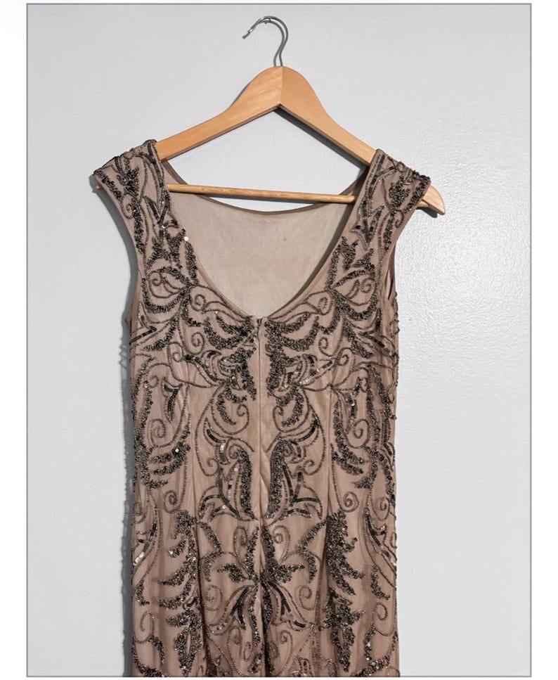 May include: A beige sleeveless dress with intricate black beadwork, hanging on a wooden hanger. The dress features a V-neck and a detailed pattern of swirling designs and embellishments, creating a vintage or formal aesthetic.