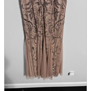 May include: A formal, floor-length dress in a blush pink hue. The dress features intricate, silver-toned beadwork in a swirling, Art Deco-inspired pattern across the bodice and skirt. The skirt has a flowing, pleated design, and the dress appears to be made of a sheer, lightweight fabric.