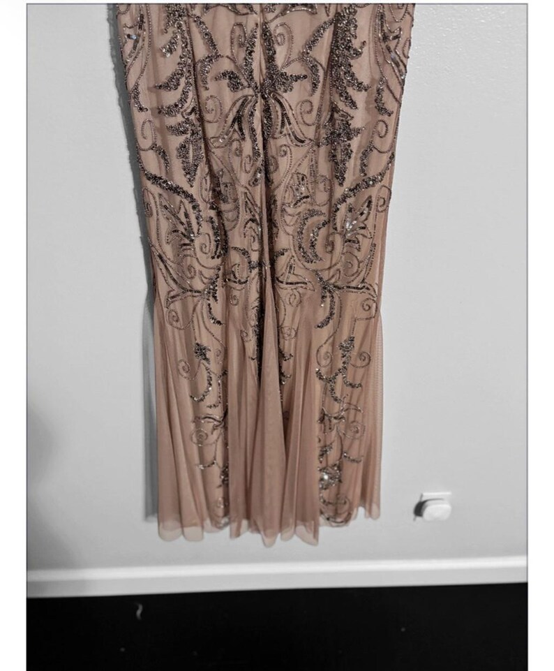 May include: A formal, floor-length dress in a blush pink hue. The dress features intricate, silver-toned beadwork in a swirling, Art Deco-inspired pattern across the bodice and skirt. The skirt has a flowing, pleated design, and the dress appears to be made of a sheer, lightweight fabric.
