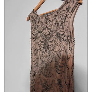 May include: A sleeveless, floor-length, champagne-colored dress with intricate dark beading and sequin embellishments. The dress is hanging on a wooden hanger. The design features a detailed, symmetrical pattern.