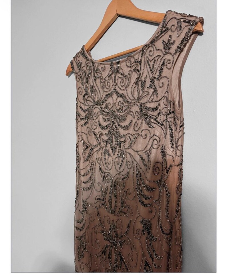 May include: A sleeveless, floor-length, champagne-colored dress with intricate dark beading and sequin embellishments. The dress is hanging on a wooden hanger. The design features a detailed, symmetrical pattern.