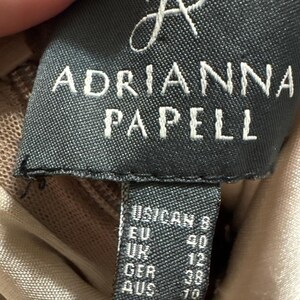 May include: Close-up of a black label with the brand name "ADRIANNA PAPELL" in white capital letters. Below the brand name are size designations for US/CAN, EU, UK, GER, and AUS.
