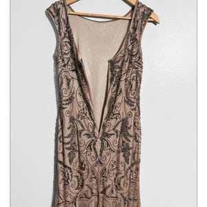 May include: A sleeveless, floor-length dress in a nude color, adorned with intricate dark beaded embellishments. The dress features a low back and a V-neckline, hanging on a wooden hanger against a white background. The beading forms a floral pattern.
