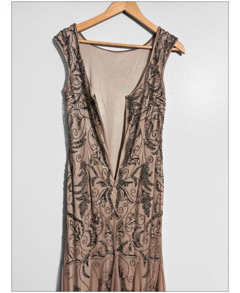 May include: A sleeveless, floor-length dress in a nude color, adorned with intricate dark beaded embellishments. The dress features a low back and a V-neckline, hanging on a wooden hanger against a white background. The beading forms a floral pattern.