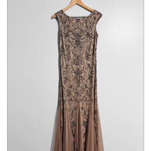 May include: A floor-length, sleeveless, beige dress with intricate black beadwork and a flowing, flared skirt. The dress is hanging on a wooden hanger against a white wall. The dress features a scoop neckline and a detailed pattern.