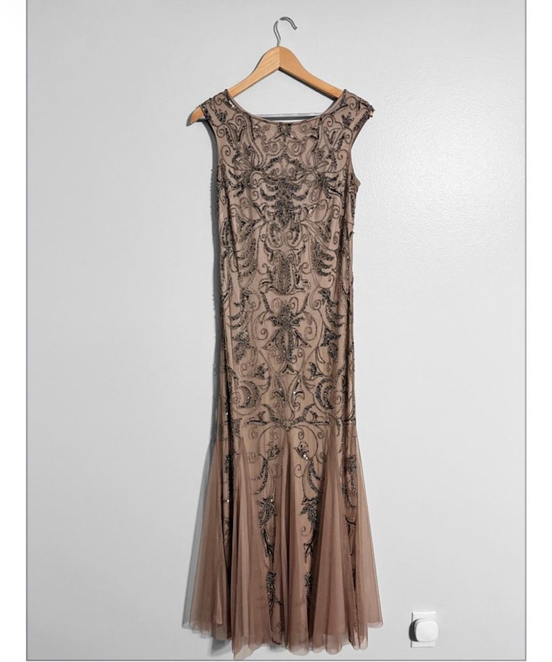 May include: A floor-length, sleeveless, beige dress with intricate black beadwork and a flowing, flared skirt. The dress is hanging on a wooden hanger against a white wall. The dress features a scoop neckline and a detailed pattern.