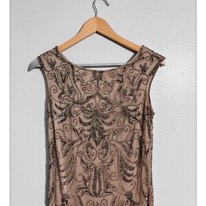 May include: A sleeveless, tan-colored dress with intricate dark beading and sequin embellishments. The dress is hanging on a wooden hanger against a plain white background. The beading forms a detailed, symmetrical pattern.