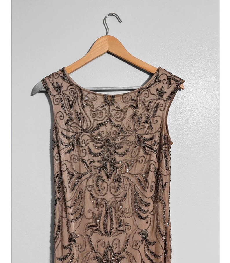 May include: A sleeveless, tan-colored dress with intricate dark beading and sequin embellishments. The dress is hanging on a wooden hanger against a plain white background. The beading forms a detailed, symmetrical pattern.