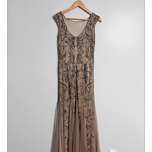 May include: A beige, sleeveless formal gown with intricate black beadwork and a flowing skirt. The dress hangs on a wooden hanger against a plain wall. The gown's design features a V-neckline and a subtle fishtail hem.