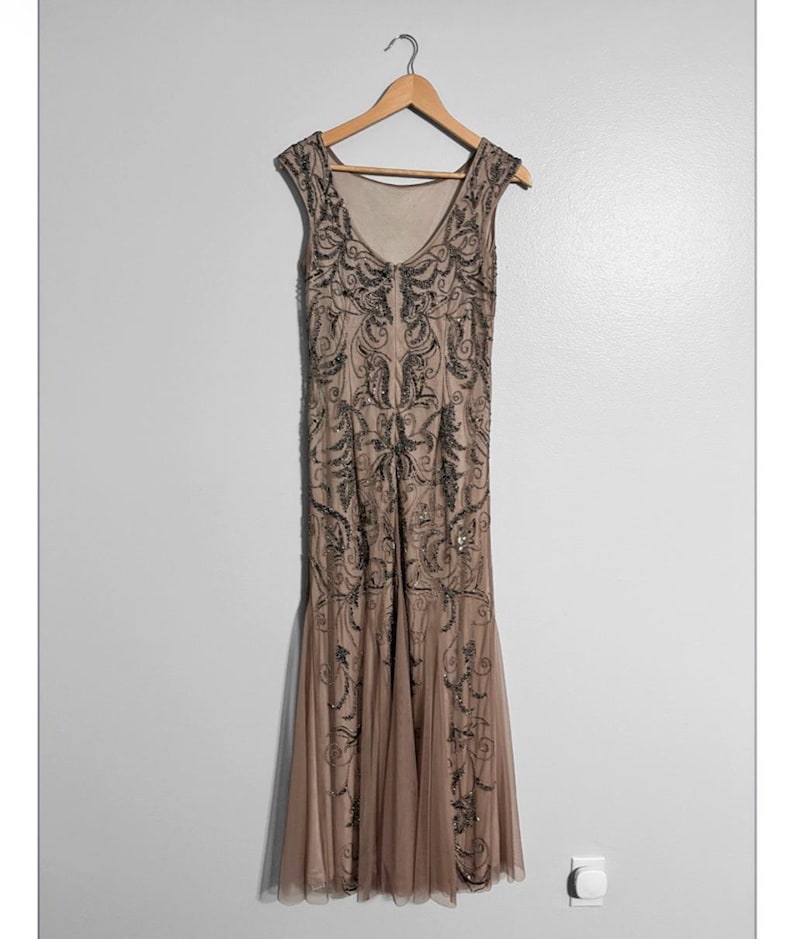 May include: A beige, sleeveless formal gown with intricate black beadwork and a flowing skirt. The dress hangs on a wooden hanger against a plain wall. The gown's design features a V-neckline and a subtle fishtail hem.