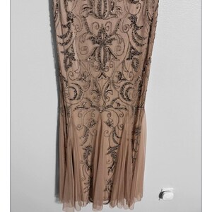 May include: A floor-length, champagne-colored dress with intricate beadwork. The dress features a fitted bodice and a flared skirt. The beadwork is in a darker, metallic tone, creating a contrast against the lighter fabric. The dress has a subtle, elegant design.