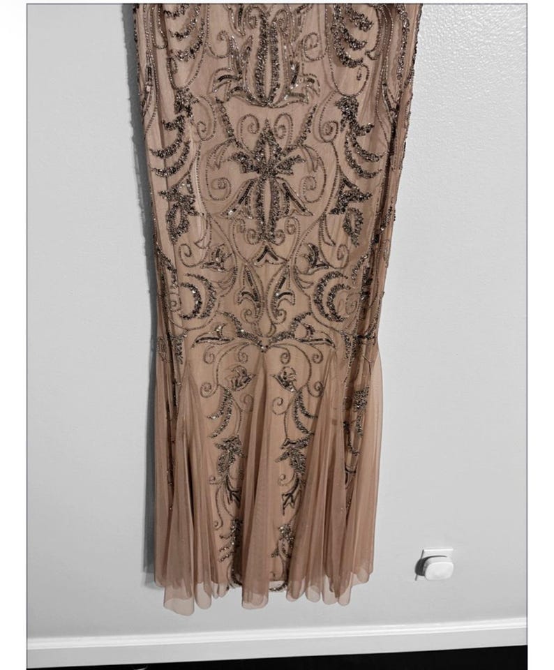 May include: A floor-length, champagne-colored dress with intricate beadwork. The dress features a fitted bodice and a flared skirt. The beadwork is in a darker, metallic tone, creating a contrast against the lighter fabric. The dress has a subtle, elegant design.