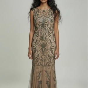 May include: A sleeveless, floor-length gown in a neutral beige hue. The dress features intricate, dark-toned embroidery in a scrolling pattern across the bodice and skirt. The skirt flares slightly at the hem, with a sheer overlay.